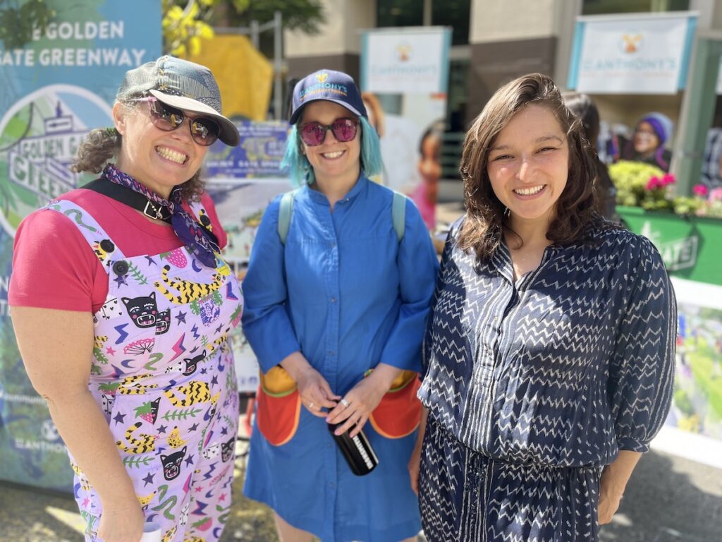 Three female volunteers pose and smile in front of the Golden Gate Greenway booth at Sunday Streets