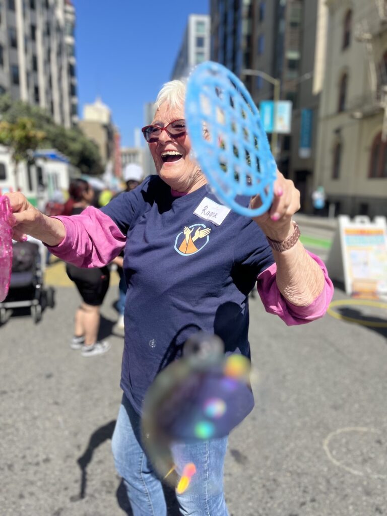 A volunteer smiles and makes a bubbles with a bubble wand at Sunday Streets on June 4th 