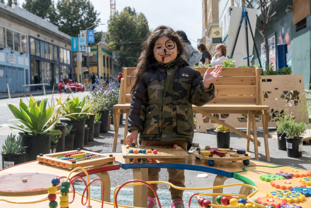 A young girl with puppy dog facepaint waves at the camera while playing with wooden toys on Golden Gate Ave during Phoenix Day in San Francisco. 